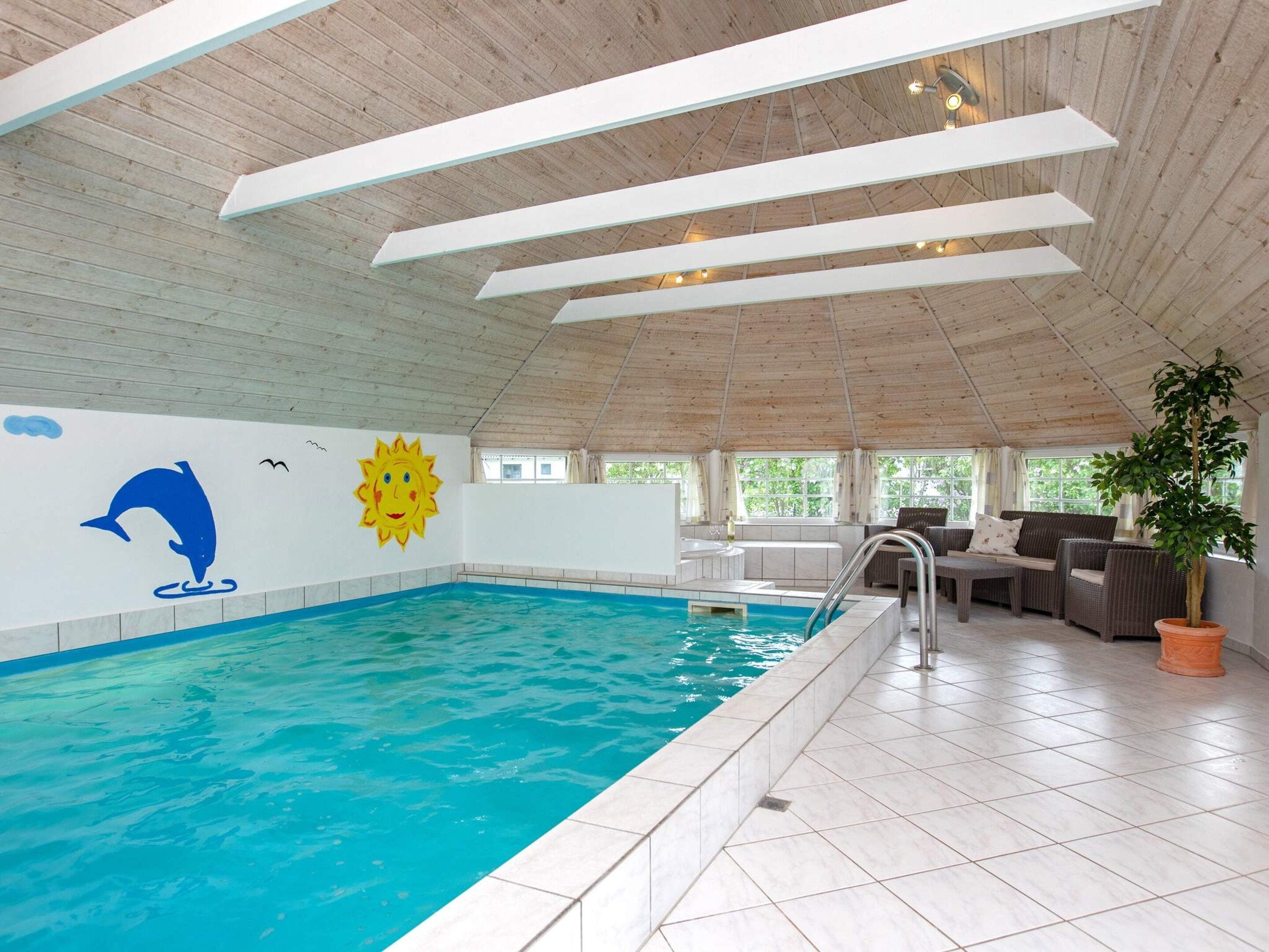 Una piscina interna con un murale a forma di delfino, un murale raffigurante il sole e una zona relax con una pianta in vaso.