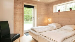 A room with wooden walls, a bed with white bedding, a black chair, and a window with plants. - Holiday Home in LÃ¦sÃ¸ (Læsø)