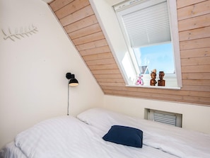 A bedroom with a sloped ceiling, a bed with white bedding, a single blue pillow, a wall-mounted lamp, and a window with blinds.