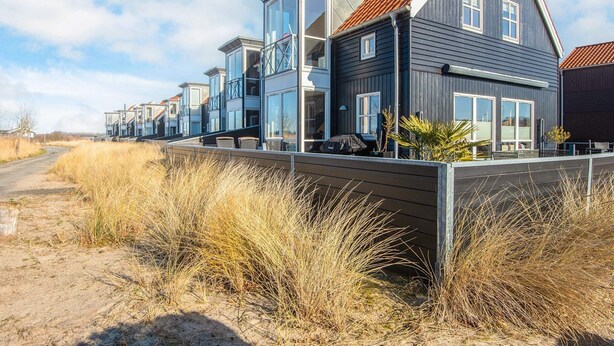 Una casa moderna frente al mar con un balcón amplio, varias ventanas y un tejado de tejas rojas.
