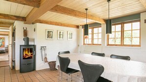 A dining area with a long table, chairs, a wood-burning stove, and large windows. - Idyllic Countryside Retreat-by Traum (Rudkobing)