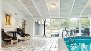 Una piscina interior con suelo de baldosas, dos sillones de descanso, una mesa y un ventanal grande.