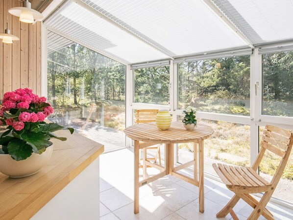A room with a wooden table and chairs, a potted plant, and a view of a forest through large windows. - Holiday Home in Strandby (Strandby)