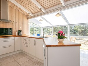 A modern kitchen with wooden ceiling beams, white cabinets, a large island with a vase of flowers, and a view of the outdoors through large windows. - Holiday Home in Strandby (Strandby)