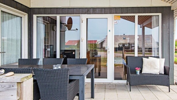 Un patio al aire libre con una mesa, sillas, un banco y un ventanal de vidrio que da a una zona techada.