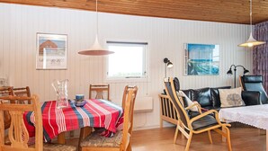Un comedor con una mesa dispuesta para cuatro personas, sillas de madera y un mantel a rayas rojas y azules. Se ve un ventanal con vistas a la vegetación, un sofá de cuero negro y una lámpara negra sobre una mesita auxiliar.