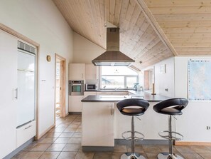 Et moderne køkken med en central ø, rustfrie hvidevarer og et højt loft med synlige bjælker.