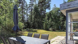 En uteplats med ett bord och stolar, omgiven av ett skogsområde och en byggnad med stora fönster.