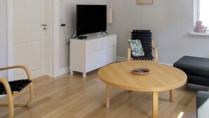 A modern living room with a round wooden table, a white cabinet with a flat-screen TV, a black chair with a green cushion, and a grey sofa.