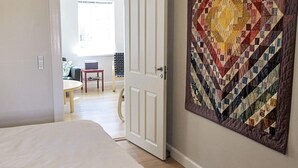 A bedroom with a large bed, a quilt on the wall, and a view into the dining area.