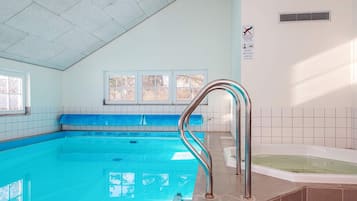 En indendørs swimmingpool med blå fliser, en ståltrappe, et spabad og vinduer med udsigt til træer.