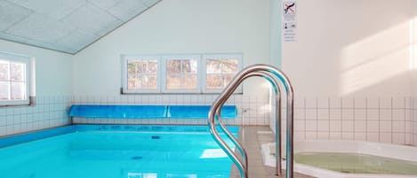 An indoor swimming pool with a blue tiled floor, a metal pool ladder, a hot tub, and windows with a view of trees.