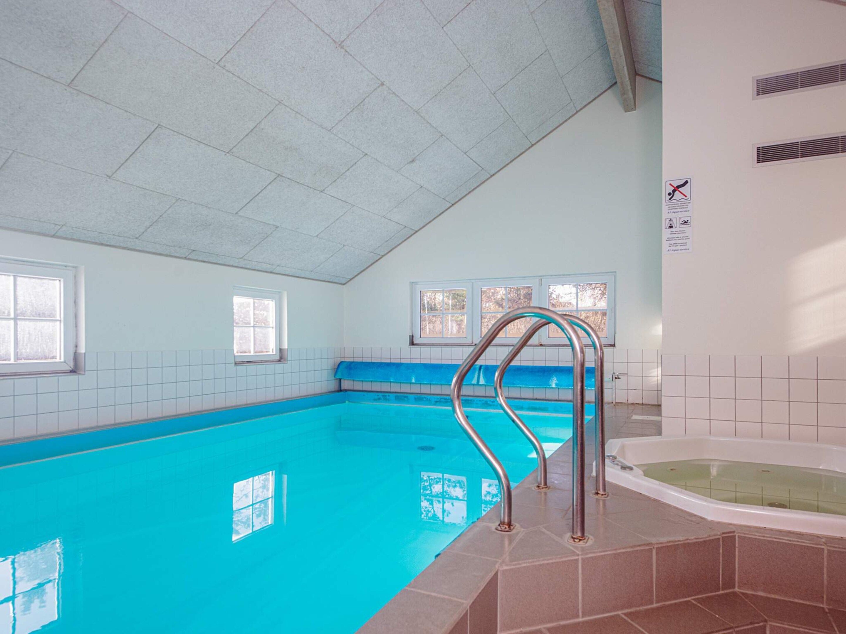 En indendørs swimmingpool med krystalklart, blåt vand, en metalstige og fliserede vægge.