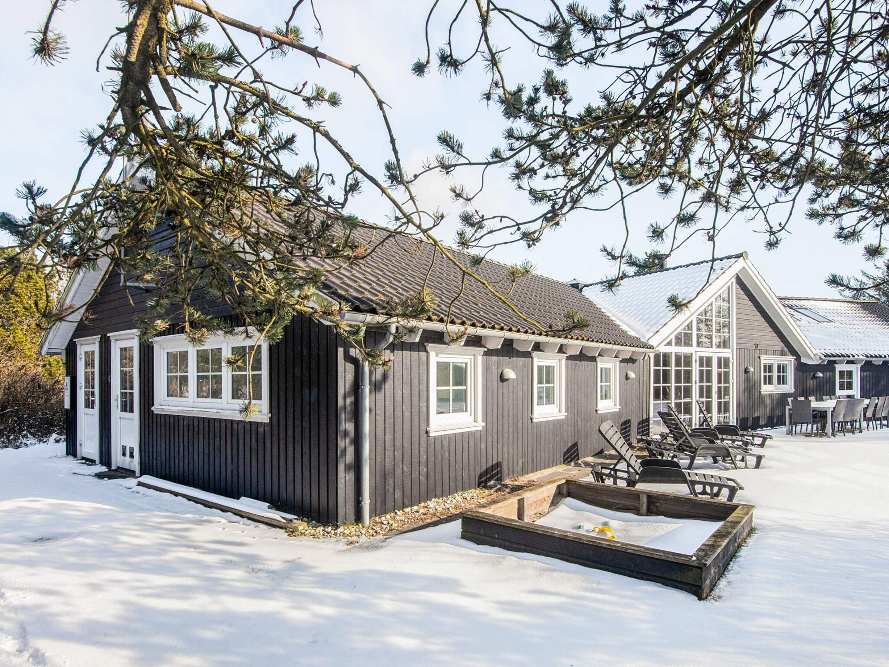 A wooden cabin with a snow-covered ground, a small outdoor seating area, and a windowed structure in the background.