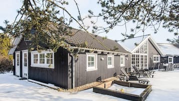 A wooden cabin with a snow-covered ground, a small outdoor seating area, and a windowed structure in the background.