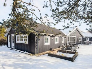 A wooden cabin with a snow-covered ground, a small outdoor seating area, and a windowed structure in the background. - 5 Star Holiday Home in Blavand (Blavand)