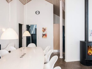 Een modern interieur met een eethoek, bestaande uit een witte tafel en stoelen, een grote hanglamp, een klok aan de muur en een houtkachel.