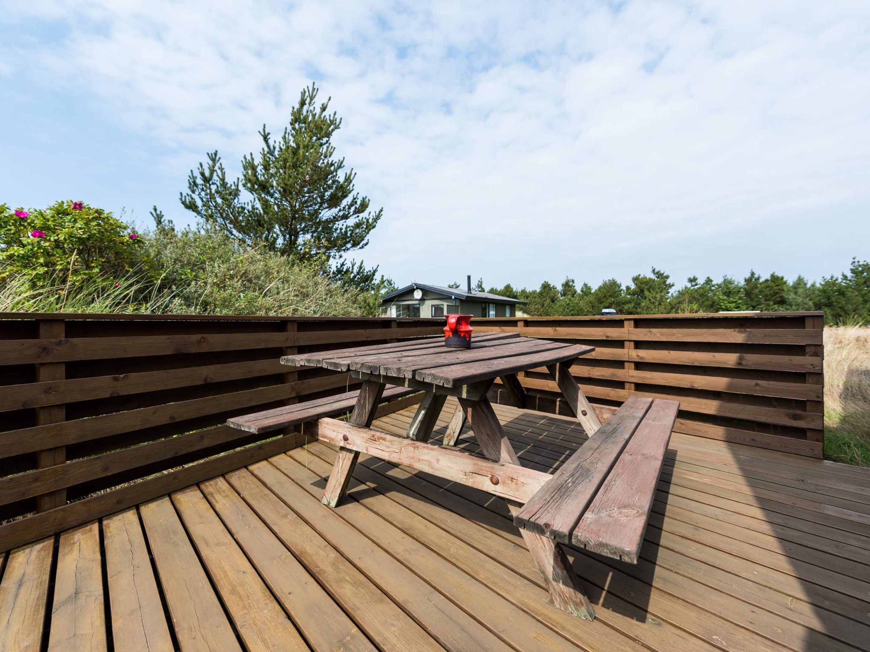 A wooden deck with a picnic table and benches, surrounded by a wooden fence and vegetation.