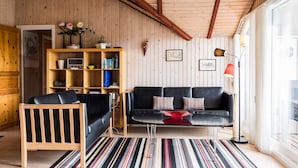 Een gezellige woonkamer met een houten plafond, een zwarte leren bank, een houten stoel, een gestreept vloerkleed en een boekenkast met diverse spullen.
