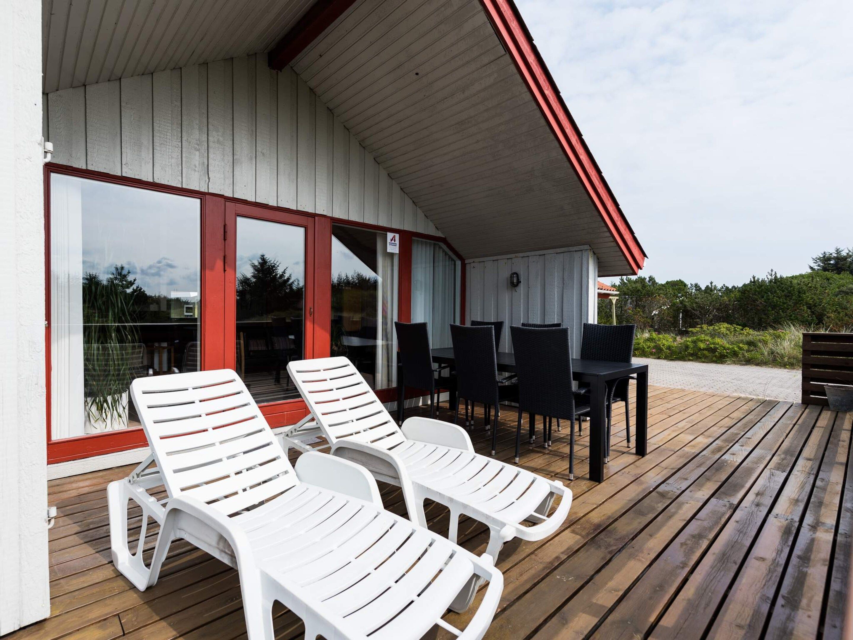 Uma área externa coberta com cadeiras de plástico brancas e uma mesa com cadeiras, um deck de madeira, e um edifício com telhado vermelho e portas de vidro.