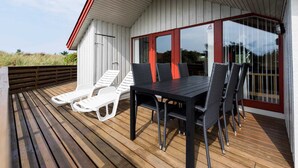 Een overdekt buitengebied met een houten terras, een tafel met stoelen en een glazen deur naar een wit gebouw met rode details.