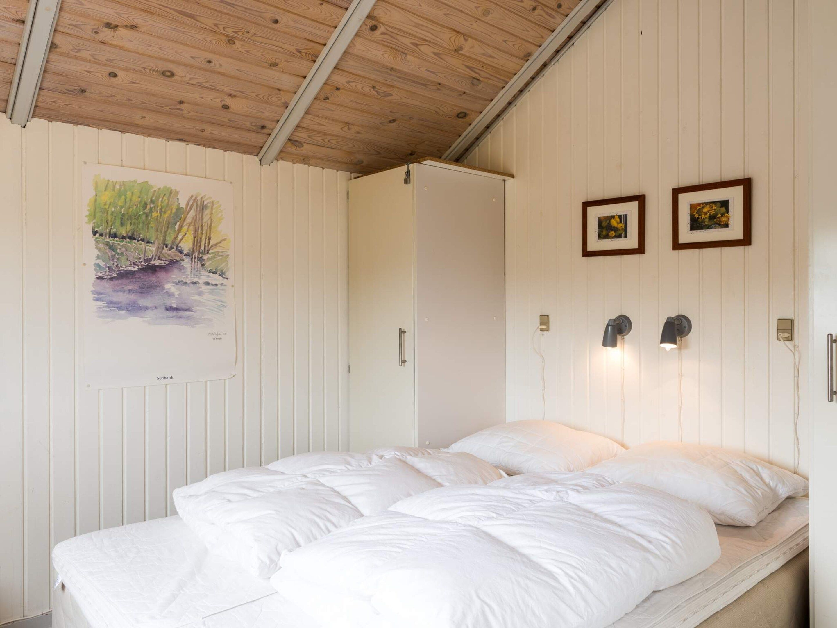 Un dormitorio con techo de madera, una cama con ropa de cama blanca, una puerta y dos luces fijadas a la pared.