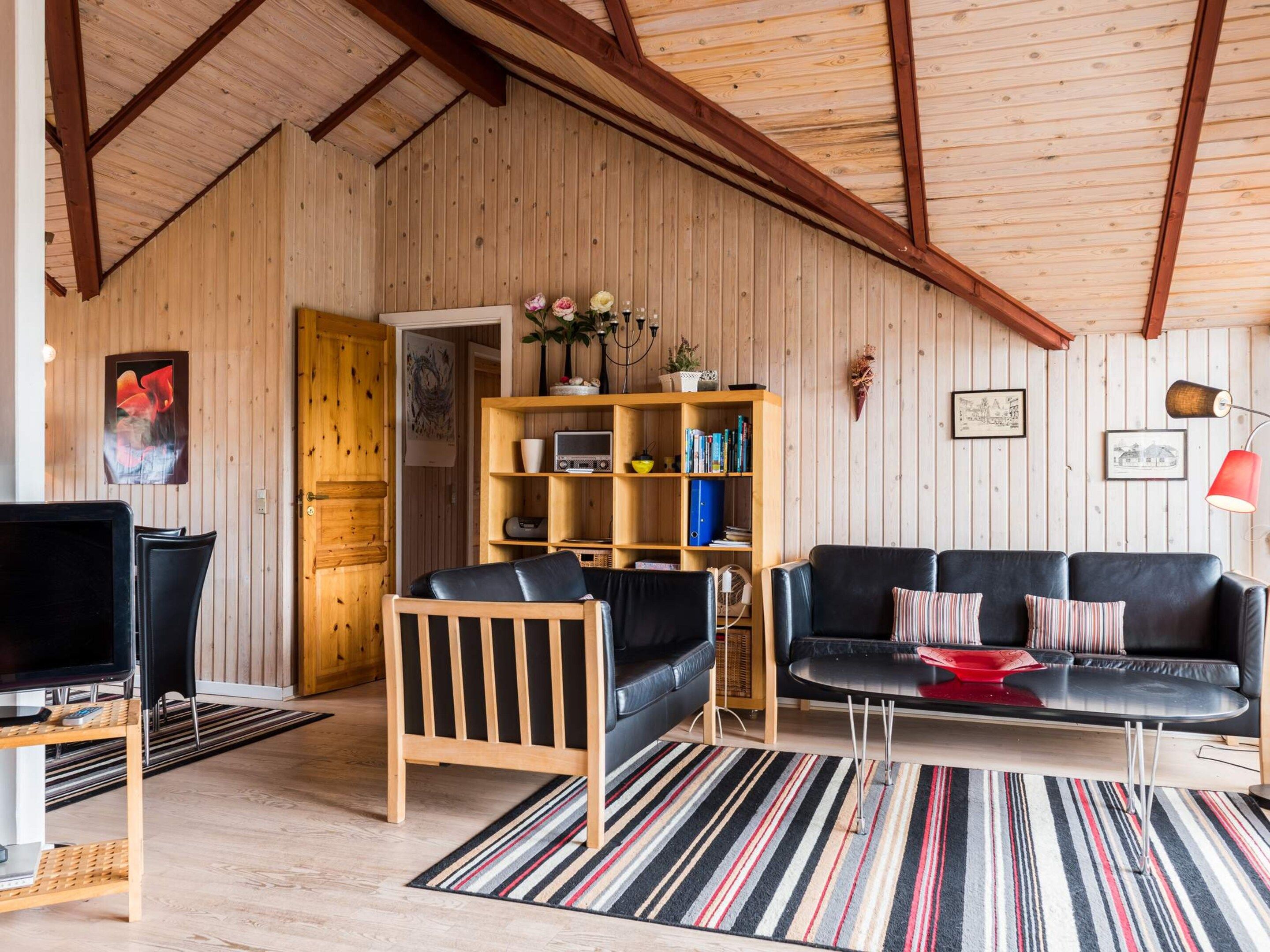 Un salón acogedor con techo de madera, un sofá de cuero negro, una silla de madera, una estantería y un televisor.