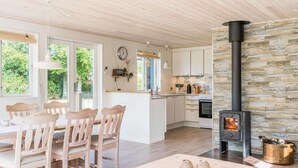 A modern kitchen with a central island, wooden chairs, a dining table, and a stone wall with a wood-burning stove. - Holiday Home in Tarm (Tarm)