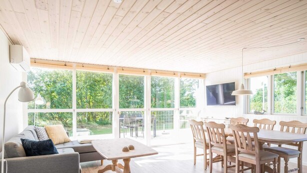 A spacious living area with a wooden ceiling, large windows, a grey sofa, a dining table with chairs, and a TV mounted on the wall. - Holiday Home in Tarm (Tarm)