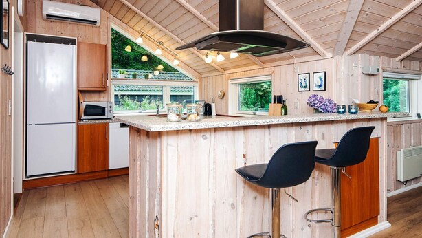 Una cocina moderna con paredes de madera, una isla central con taburetes de bar, una campana extractora de acero inoxidable y vistas al exterior con vegetación.