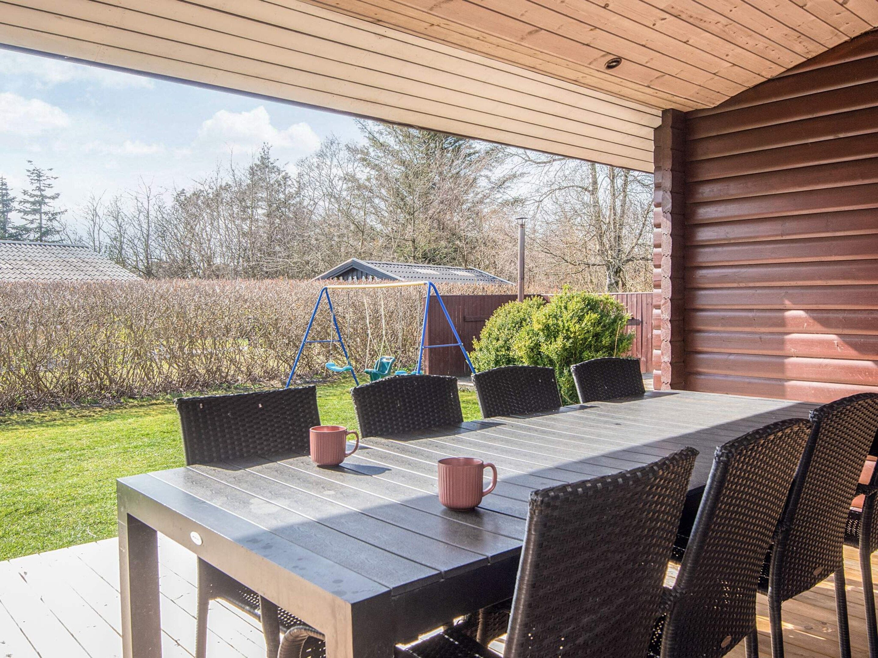 An outdoor patio with a wooden table and chairs, a swing set, and a covered area with a view of a backyard and trees.