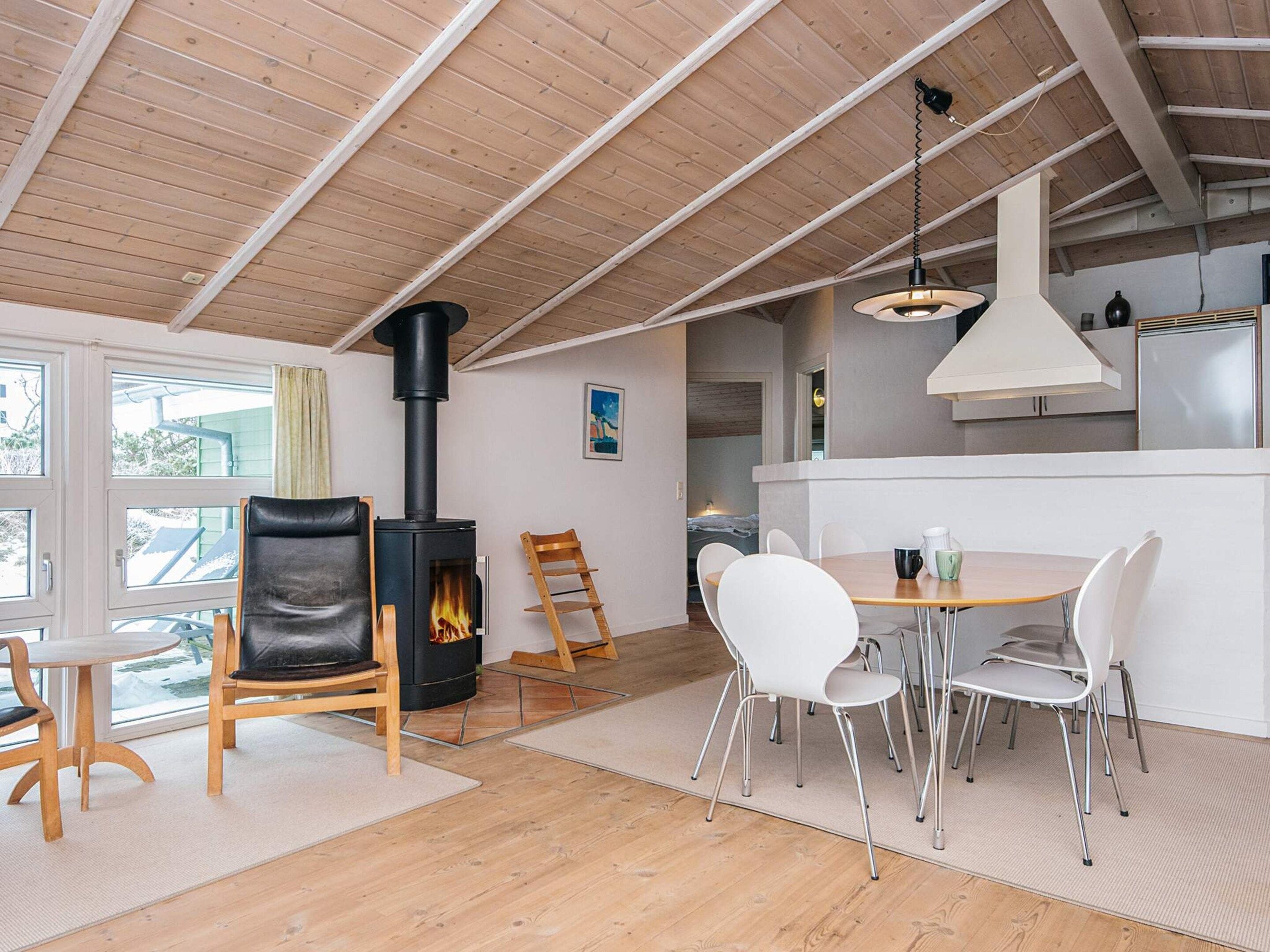 Een modern interieur met een houtkachel, een eethoek met witte stoelen, en een keuken met roestvrijstalen apparatuur.