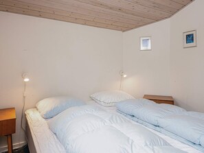 Een slaapkamer met twee bedden, elk met een houten hoofdbord en witte beddengoed. Er zijn twee nachtkastjes, elk met een lamp. De kamer heeft witte muren en een houten plafond.