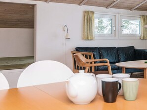 A cozy room with a wooden ceiling, a black leather sofa, a wooden chair, a small table with a white pitcher and mugs, and a window with curtains. - 6 Person Holiday Home in Blavand (Blavand)