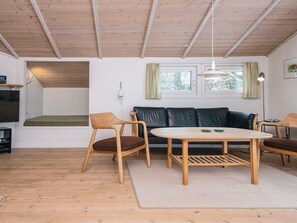 Un salon chaleureux avec un plafond en bois, un canapé en cuir noir, des chaises en bois, une table ronde en bois et un téléviseur placé à gauche.