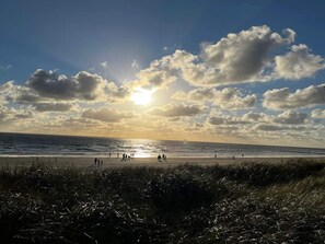 Een strand met mensen, gras op de voorgrond en een ondergaande zon.