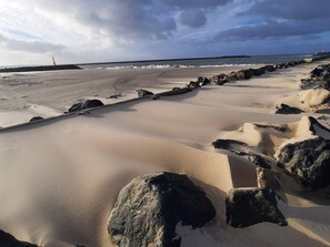 A beach with sand dunes and rocks, a lighthouse in the distance, and a cloudy sky.