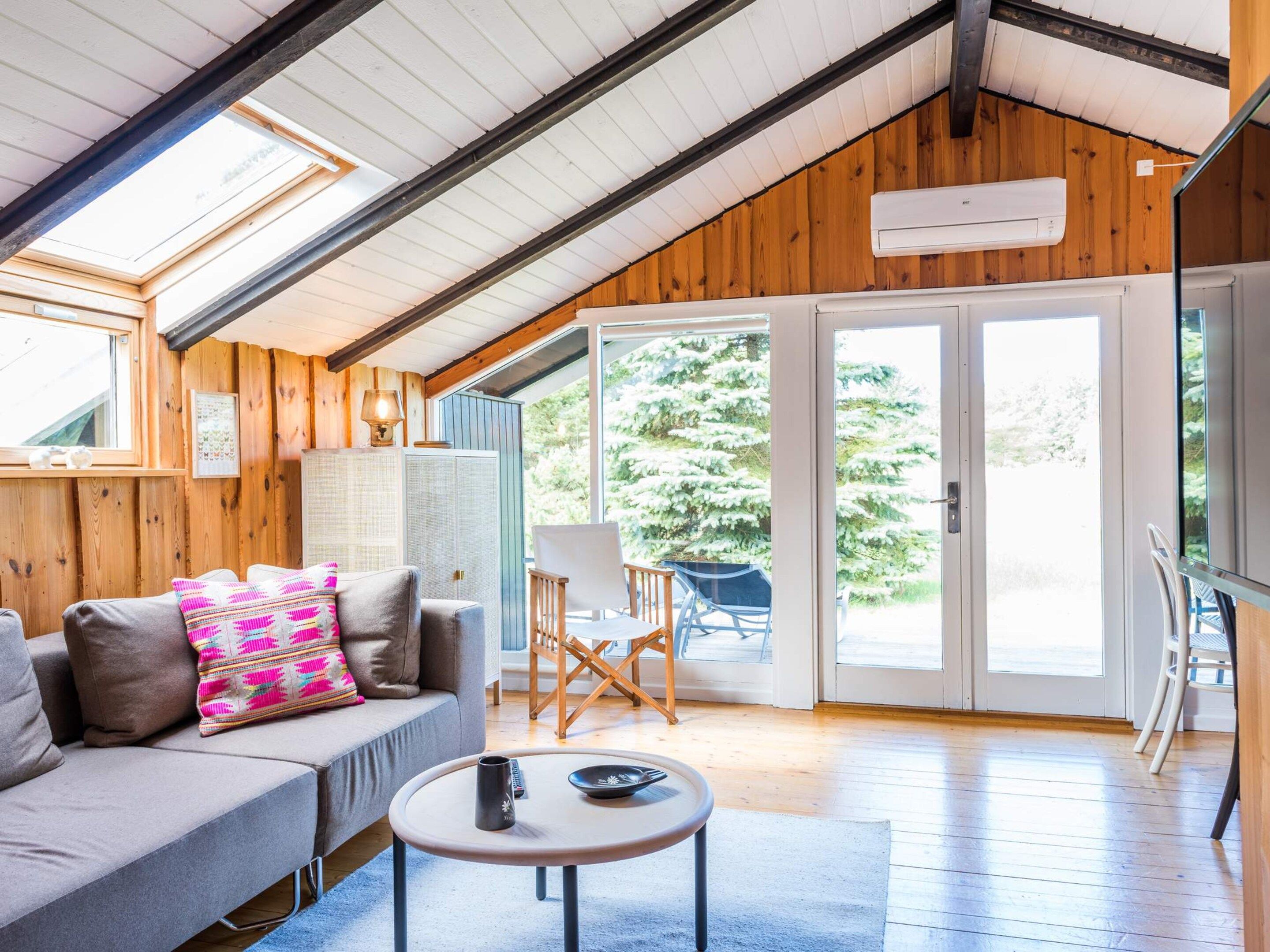 Ein gemütliches Zimmer mit schrägen Wänden, Holzwänden, einer Couch mit bunten Kissen, einem kleinen runden Tisch und Blick auf eine verschneite Landschaft durch eine Glastür.