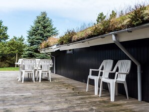 Un espace extérieur abrité doté d’une terrasse en bois, de chaises en plastique blanches et d’un bâtiment noir surmonté d’un toit vert.