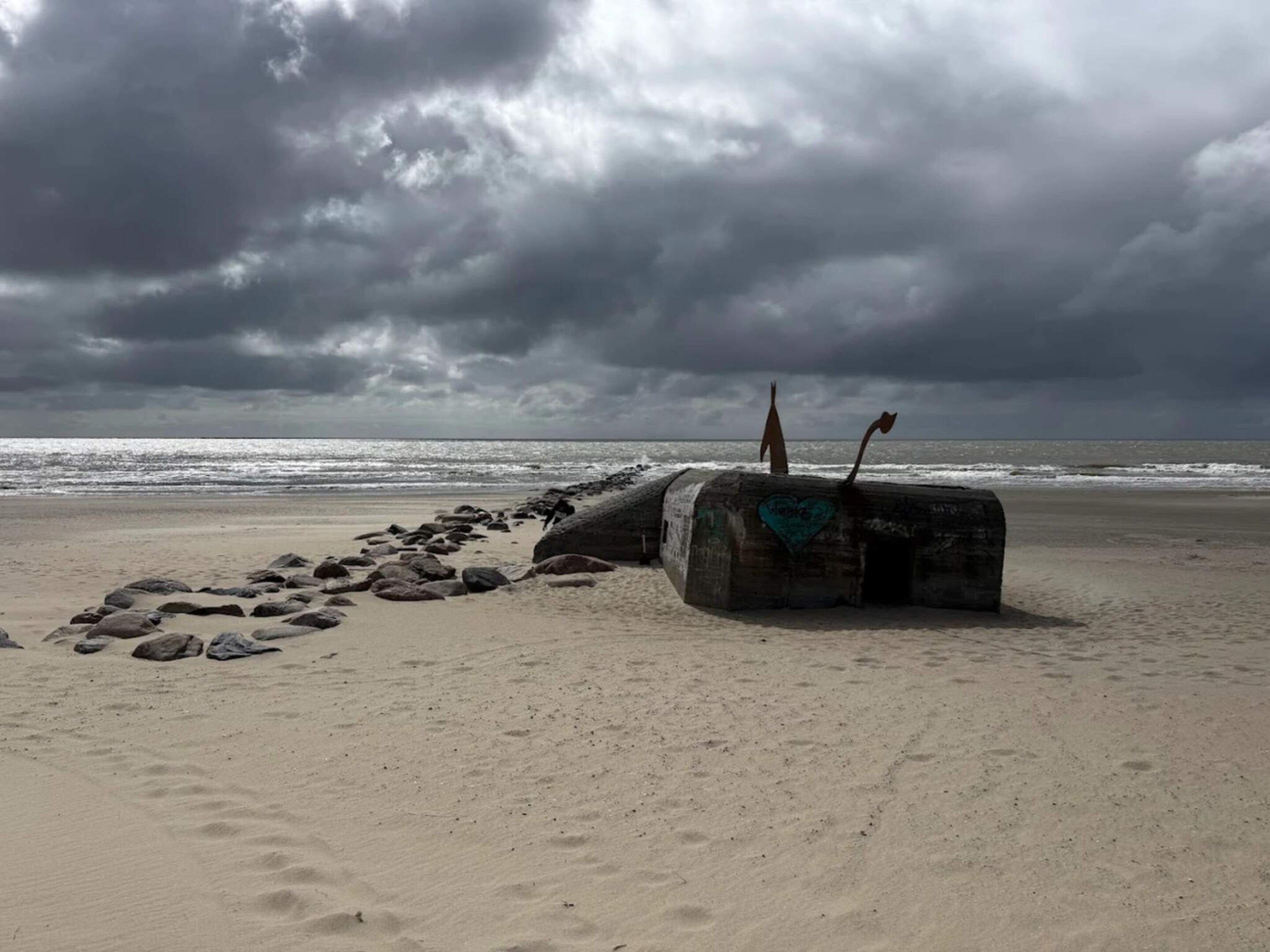 A beach with a large log and two metal sculptures, a sandy path leading to the water, and a cloudy sky.