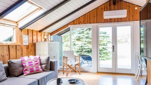A cozy room with a sloped ceiling, wooden walls, a sofa with colorful pillows, a small table with a remote control, and a view of a forest through large glass doors. - 6 Person Holiday Home in Blavand (Blavand)
