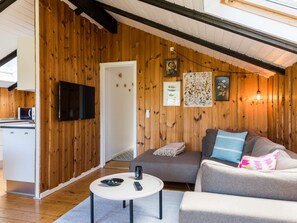 A cozy living area with a wooden interior, a flat-screen TV mounted on the wall, a sofa with cushions, a small round table with a remote control, and a door leading to another room.
