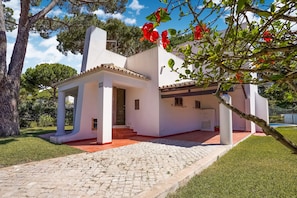 A white building with a tiled roof, a paved entrance, and a garden area with a tree and red flowers. - Villa in Vilamoura With Private Pool (Loulé)