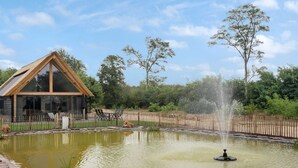 Una cabaña de madera con ventanas amplias, un estanque con una fuente y un jardín vallado.