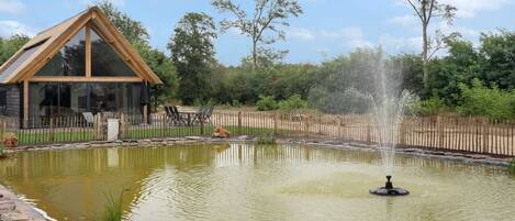 A wooden cabin with large windows, a pond with a fountain, and a fenced garden area.