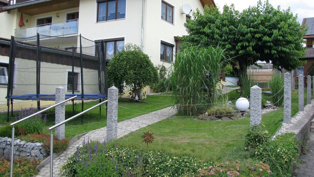 A residential building with a balcony, a garden with various plants, a trampoline, and a stone pathway. - Ferienwohnung im Bayerischen Wald mit Balkon (Drachselsried)