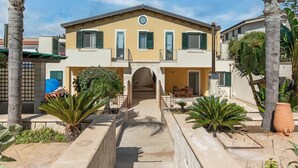 A two-story building with a central entrance, a balcony on the upper floor, and a paved courtyard with palm trees and planters. - Simplistic Apartment in Reitani near Lido Beach (Noto)