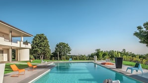 A modern outdoor swimming pool with lounge chairs and a table, surrounded by greenery and a building with balconies. - Holiday Home Near Urbino & Beaches (Tavullia)