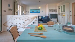A cozy living room with a sofa, a dining table set with plates and cutlery, a hanging net with shells, and a painting of a coastal scene. - Holiday Home Near Urbino & Beaches (Tavullia)