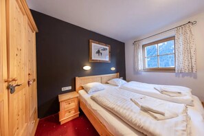 A bedroom with two beds, a wooden wardrobe, a small bedside table, a wall-mounted light, and a window with curtains.
